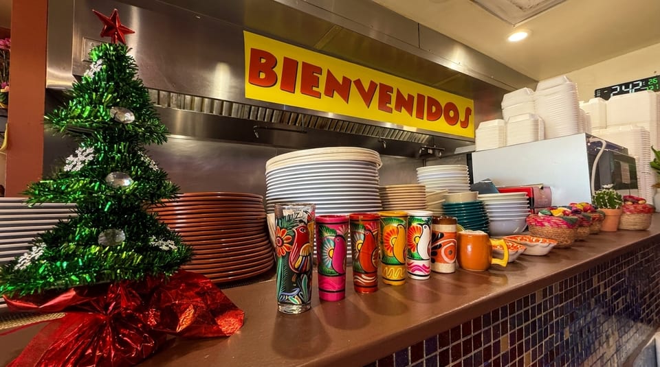 A small Christmas tree and colorful Mexican-themed glasses on a restaurant counter with a "BIENVENIDOS" sign.