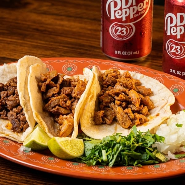 Three tacos with meat, lime, cilantro, and onions on an orange plate, with two Dr. Pepper cans.