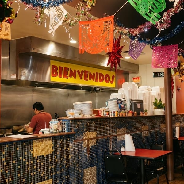 A vibrant Mexican restaurant interior with colorful papel picado, a "Bienvenidos" sign, and a chef cooking.