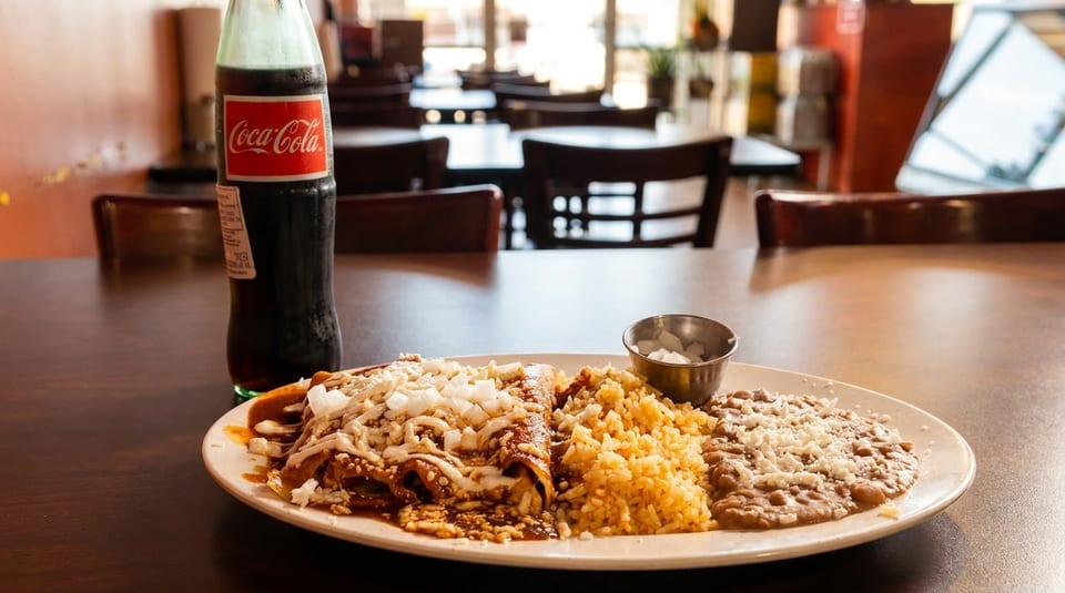A plate of enchiladas with rice and beans, and a glass bottle of Coca-Cola on a restaurant table.