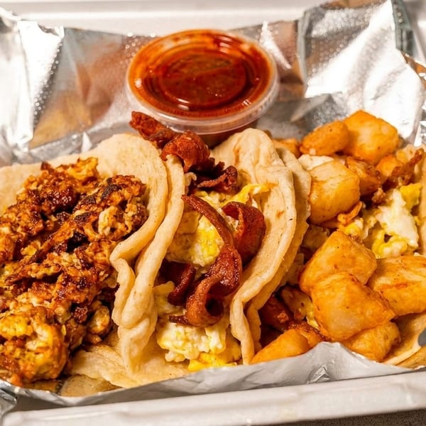 Three breakfast tacos with eggs, bacon, and potatoes, served with salsa in a to-go container.