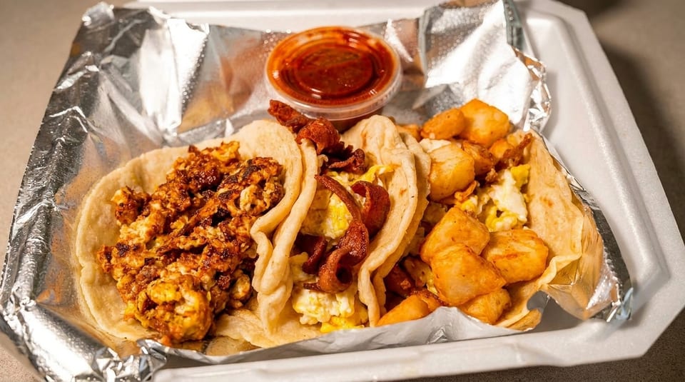 Three breakfast tacos with eggs, bacon, and potatoes, served with salsa in a to-go container.