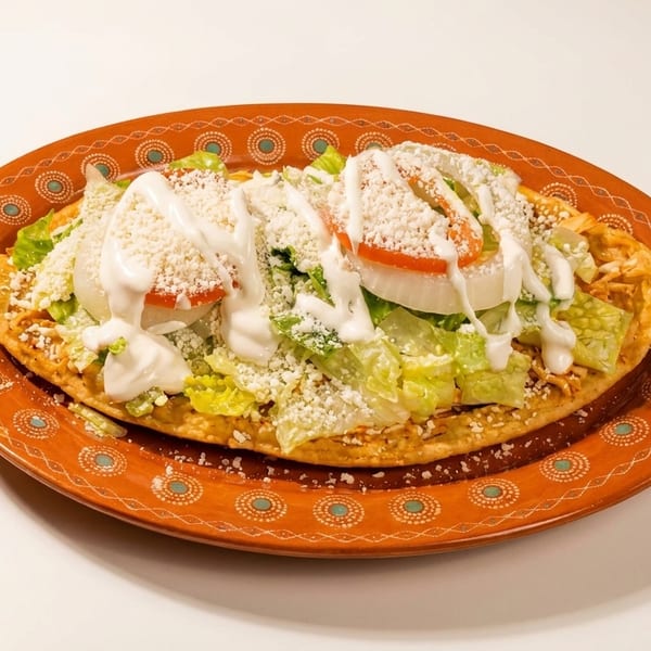 A large tostada with shredded chicken, lettuce, tomato, onion, sour cream, and crumbled cheese.